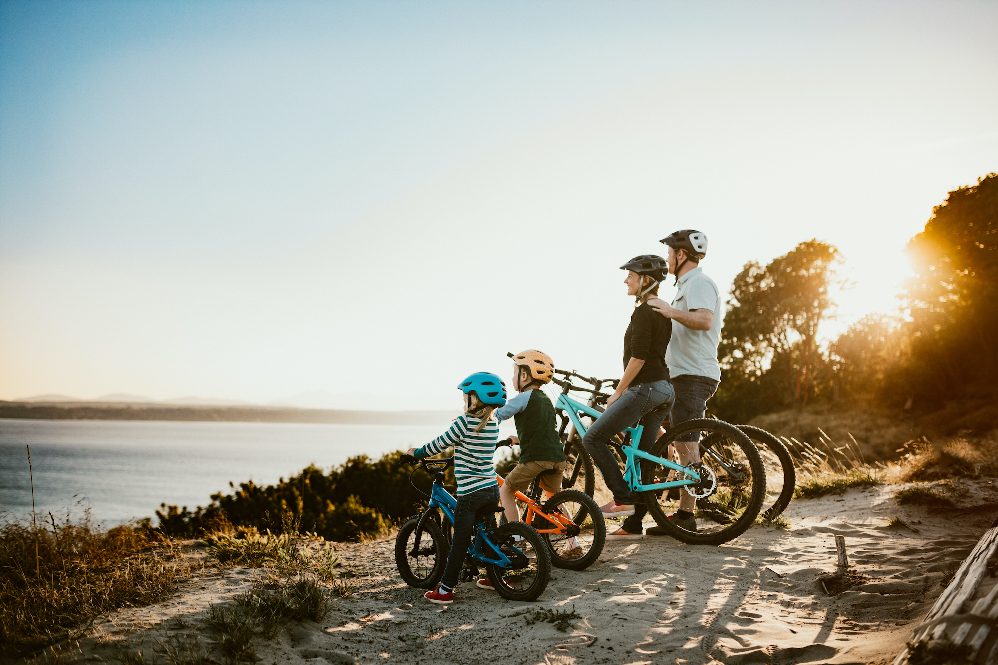 A father and mother ride mountain bikes together with their two small children.  A fun way to spend time together and exercise while on vacation in the Seattle, Washington area.  They stop and look at a view of the Puget Sound from a ridge top.